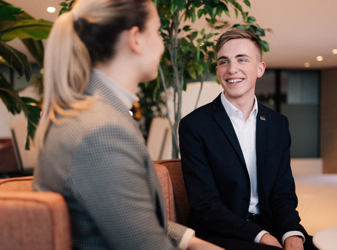 Junge Frau und junger Marriott Auszubildender Mann in Businesskleidung sitzen und unterhalten sich lächelnd in einer Hotellobby