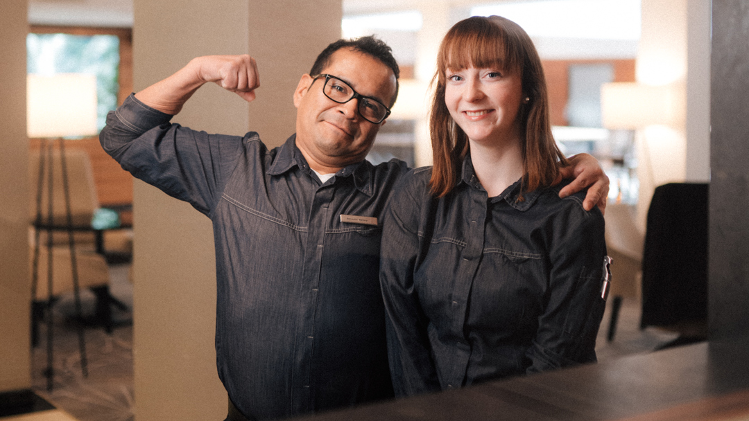 Zwei Marriott Hotelmitarbeiter, ein Mann mittleren Alters mit Brille und junge Frau mit roten Haaren stehen lächelnd nebeneinander. Der Mann legt den Arm um die Frau und hebt den zweiten Arm zu einer Siegerpose