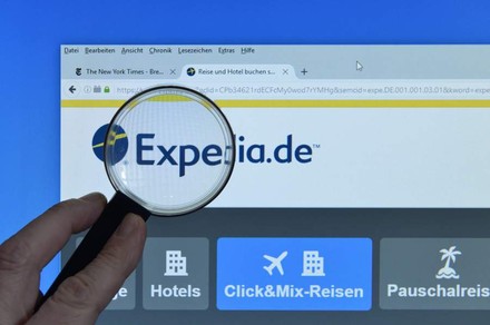 Mann hält Lupe vor Logo von Expedia.de an Bildschirm