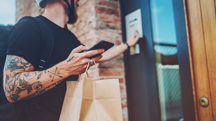 Junger Mann mit Motorradhelm hält ein Smartphone und eine Lieferservice-Tüte in der Hand und klingelt an einer Tür