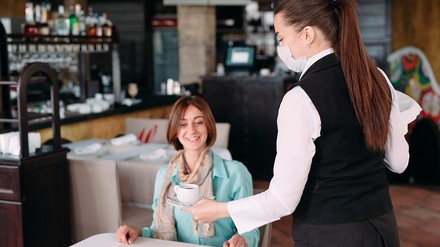 Bedienung in einem Restaurant oder Cafe trägt eine Mund-Nasen-Maske, während sie einen Gast bedient