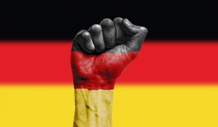 Eine Hand zur Faust geballt in Deutschlandfarben angemalt, im Hintergrund die Deutschlandflagge