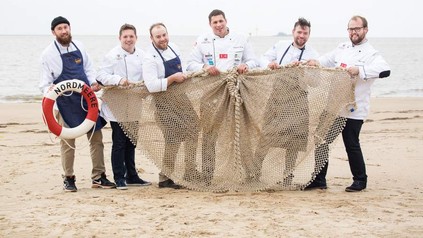 Sechs Köche halten gemeinsam ein Fischernetz an einem Strand