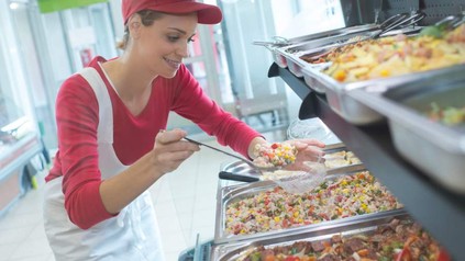 Eine junge Kantinenkraft löffelt Salat aus einer großen Edelstahlwanne in eine Plastikschüssel