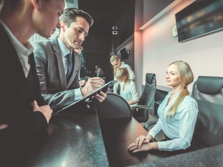 Zwei Geschäftsleute stehen mit einem Klemmbrett vor einer vollbesetzten Hotelrezeption und besprechen etwas mit der Rezeptionistin