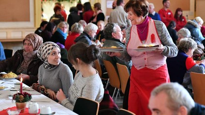 Eine ältere Dame serviert einigen Personen an einem langen Tisch Speisen, der Saal ist gut besucht
