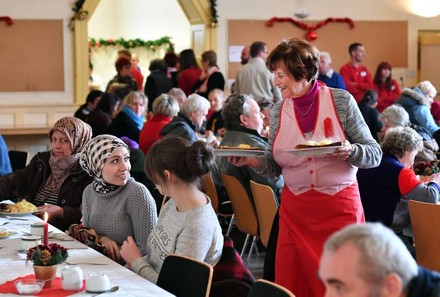 Eine ältere Dame serviert einigen Personen an einem langen Tisch Speisen, der Saal ist gut besucht