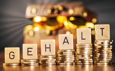 Das Wort Gehalt auf ansteigenden Stapeln mit Münzen vor einer Truhe mit Geld im Hintergrund.