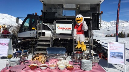 Ein kleines Buffett ist vor einer Schneeraupe aufgebaut auf dem ein Schild Grächen St. Niklaus steht. Auf den Stufen zur Raupe steht eine Person als Maskottchen in einem Vogelkostüm verkleidet