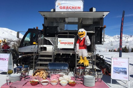 Ein kleines Buffett ist vor einer Schneeraupe aufgebaut auf dem ein Schild Grächen St. Niklaus steht. Auf den Stufen zur Raupe steht eine Person als Maskottchen in einem Vogelkostüm verkleidet