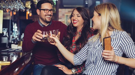 Drei Freunde sitzen lachend an einer Bar-Theke und stoßen mit Drinks an