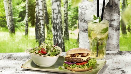 Eine Portion Burger und Salat mit einem frischen Getränk im Restaurant Hans im Glück. Im Hintergrund sieht man Birkenbäume