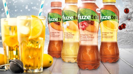 Vier verschiedene Flaschen fuzetea, Schwarzer Tee Pfirsich, Grüner Tee Mango Kamille, Schwarzer Tee Pfirsich Hibiskus und Schwarzer Tee Zitrone, davor stehen zwei Gläser mit Eistee, Eiswürfen und Strohhalm