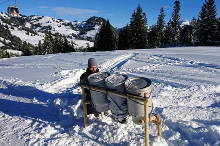 Ein Mann kniet im Schnee vor drei großen Milchkannen, die dort aufgestellt wurden