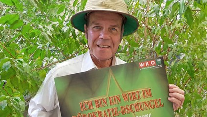 Peter Dobcak hält ein Schild auf dem steht Ich bin ein Wirt im Bürokratie-Dschungel Holt mich hier Raus!