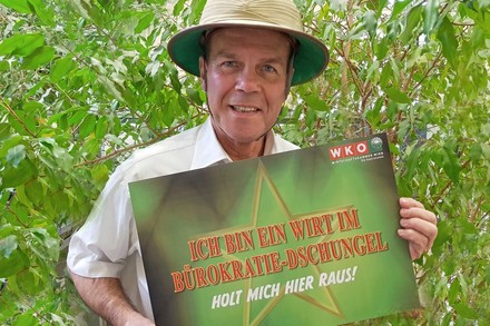 Peter Dobcak hält ein Schild auf dem steht Ich bin ein Wirt im Bürokratie-Dschungel Holt mich hier Raus!