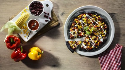 Eine Pizza aus schwarzem Teig mit Mais, Paprika und Kidney-Bohnen belegt, daneben die Zutaten des Belages