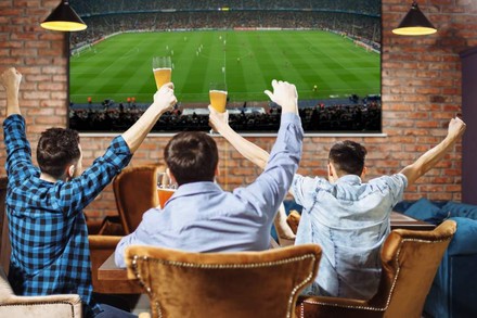 Drei Junge Männer jubeln mit Bier in der Hand vor einem großen Fernseher auf dem ein Fußballspiel läuft