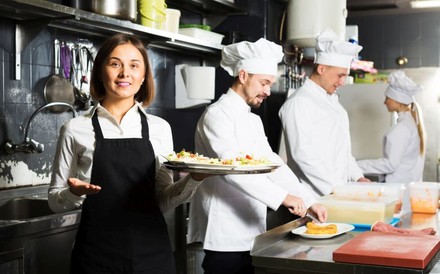Kellnerin nimmt volle Teller in der Küche zum Servieren auf. Im Hintergrund 2 Köche.