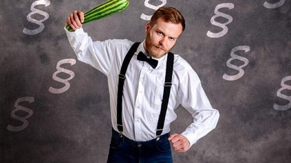 Eine junger Mann schwingt einen Zucchini wie einen Knüppel, im Hintergrund sind Paragraphenzeichen zu sehen