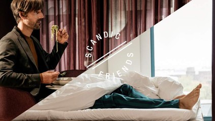 Ein Mann sitzt an einem Tisch beim Essen, seine Beine jedoch befinden sich bereits im Bett. Davor steht Scandic Friends