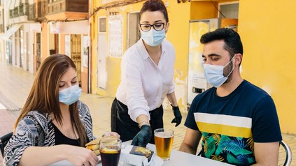 Kellnerin mit Maske bedient ein Paar an einem Tisch, beide ebenfalls mit Maske