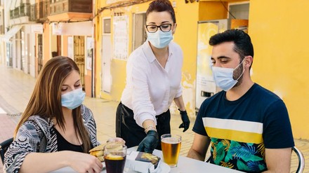 Kellnerin mit Maske bedient ein Paar an einem Tisch, beide ebenfalls mit Maske