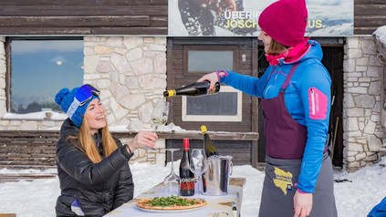 Eine junge Servicekraft schenkt einer Wintersportlerin auf einer Hütte im Schnee Wein nach