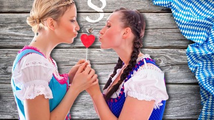 Zwei Frauen im Dirndl halten einen herzförmigen Lolli zwischen sich um ihn zu küssen, darüber ist ein Paragraphenzeichen abgebildet