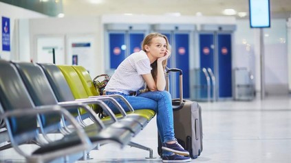 Eine junge Frau sitzt mit ihrem Koffer gelangweilt in einer Wartehalle am Flughafen