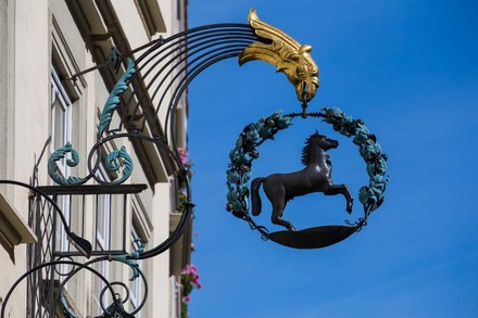 Ein Eisengestell das ein Pferd in einem Lorbeerkranz, gehalten von einem goldenen Vogelkopf zeigt hängt an einer Hauswand