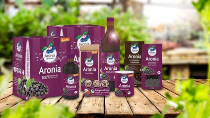 Diverse Produkte mit Aronia zubereitet, zum Beispiel in Schokolade, getrocknete Aronia, Früchtetee oder Saft von demeter auf einem Holztisch