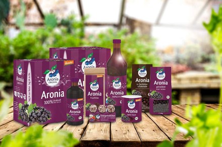 Diverse Produkte mit Aronia zubereitet, zum Beispiel in Schokolade, getrocknete Aronia, Früchtetee oder Saft von demeter auf einem Holztisch
