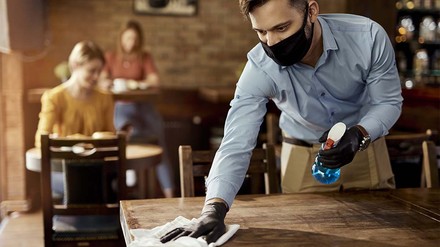 Junge Servicekraft mit medizinischer Maske putzt einen Tisch im Restaurant