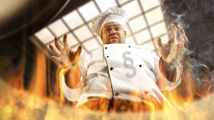 Ein verzweifelter Koch blickt in einen brennenden Topf. Auf seiner Kochjacke ist ein Paragraphenzeichen zu sehen