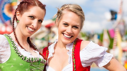 Zwei junge Frauen im Dirndl stehen auf der Theresienwiese. Im Hintergrund sind Fahrgeschäfte.