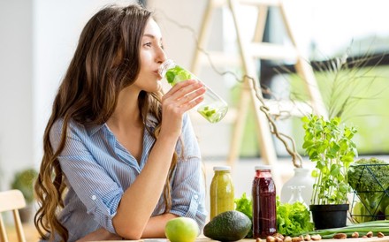Eine frau sitzt an einem Tisch mit Smoothies, einer Avocado und einem Apfel und trinkt aus einer Flasche mit Wasser und Kräutern