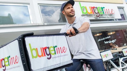 Lieferfahrer mit weißem Shirt und Cap auf Fahrrad mit Lieferbox von Burgerme