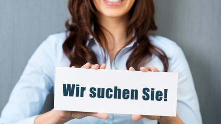 Eine Frau hält ein Schild vor sich auf dem „Wir suchen Sie“ steht