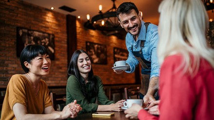 Mann bedient drei junge Frauen an einem Tisch in einem Cafe