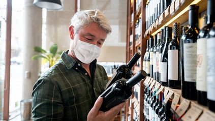 Älterer Mann mit Mund-Nasen-Schutz steht mit zwei Weinflaschen in den Händen vor einem Weinregal und liest die Etiketten