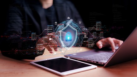 Mann sitzt vor Laptop und Tablet. Zeigt mit Finger auf Sicherheits-Symbol.