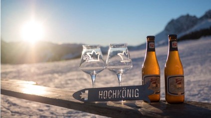 Zwei Flaschen Craft Bier und dazugehörige Gläser in einer Schneelandschaft mit einem Schild auf dem Hochkönig steht