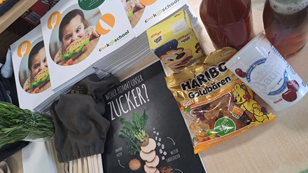 Auf einem Tisch liegt ein Kochbuch, ein Informationsblatt zu Zucker, Gummibären und ein Joghurtbecher.