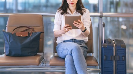 Frau sitzt mit Koffer und Tablet in der Hand am Flughafen