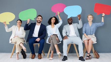Fünf Mitarbeiter sitzen auf Stühlen und heben bunte Sprechblasen in die Luft