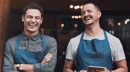 Zwei Mitarbeiter aus der Gastronomie stehen lachend im Eingangsbereich eines Betriebs und tragen Arbeitsschürzen