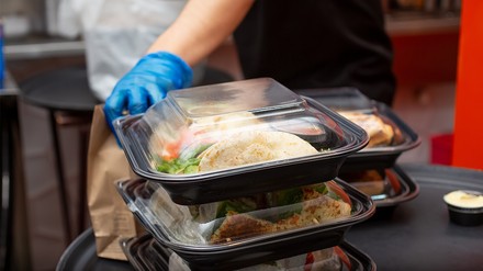 Mitarbeiter stapelt To-go-Boxen mit Essen zum Mitnehmen auf einem Tisch, trägt blaue Einweghandschuhe.