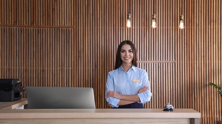 Hotelrezeption mit Mitarbeiterin am Empfang; Schreibtisch mit Laptop, Klingel und warmen Hängelampen vor Holzlamellenwand.