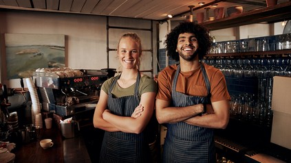 Zwei Mitarbeiter stehen lächelnd hinter der Theke eines Cafés und tragen Schürzen; im Hintergrund sind Kaffeemaschine und Gläserregal zu sehen.